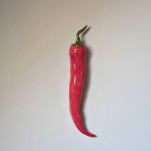 Könnte beinhalten: Eine glänzende rote Chilischoten-Skulptur mit einem glatten, geschwungenen Körper und einem kleinen, spitzen grünen Stiel an der Oberseite. Die Oberfläche der Chilischote weist subtile Variationen in Rottönen auf, was dem Objekt ein realistisches Aussehen verleiht.