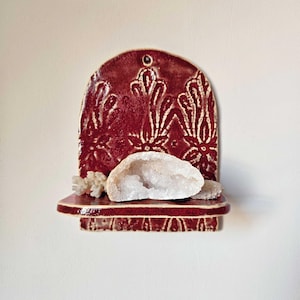 Könnte beinhalten: Ein rotes Keramikregal mit einem dekorativen Blumendesign und einer weißen Geode. Das Regal ist an einer weißen Wand befestigt. Das Regal ist etwa 15 cm breit.