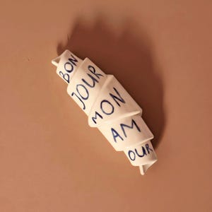 Op de afbeelding: Witte keramische croissant sculptuur met blauwe tekst "BON JOUR MON AMOUR". De sculptuur staat op een lichtbruin oppervlak. De croissant is een decoratief item.