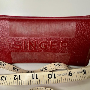 Puede incluir: Un estuche rojo para máquina de coser Singer con el nombre de la marca en relieve en la parte delantera. Una cinta métrica vintage con números y marcas negras está envuelta alrededor del estuche. El estuche es rectangular con una superficie texturizada.
