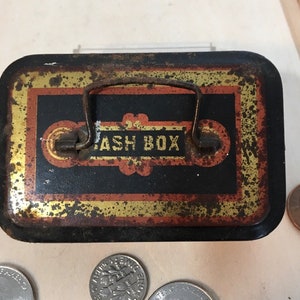 Könnte beinhalten: Vintage Metall-Geldkassette mit rostiger, alternder Oberfläche. Die Kasse hat einen schwarzen Hintergrund mit goldenen und roten Akzenten. Der Deckel verfügt über einen Metallgriff und die Aufschrift "CASH BOX" in goldenen Buchstaben.