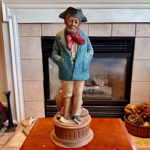 May include: A figurine of a man wearing a blue coat, a red scarf, and a black hat. He is standing on a wooden base with a barrel at his feet.