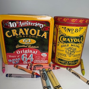 Puede incluir: Una caja de crayones Crayola amarilla del 40 aniversario con 64 unidades y una lata de crayones Crayola n.º 8 roja y amarilla. Varios crayones de varios colores están esparcidos alrededor de las cajas. Las cajas y los crayones son para el trabajo de color educativo.