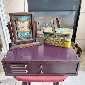May include: A dark wood swivel clock with a square face, a rectangular metal lunchbox featuring a colorful outdoor scene with figures, and a purple two-drawer metal storage cabinet with brass-toned label holders and small round knobs.