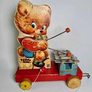 May include: Vintage "Teddy Zilo" pull-along toy featuring a bear playing a xylophone. The bear is brown with blue eyes, wearing a red vest and holding mallets. The xylophone has silver bars and orange beaters, mounted on a red wooden base with wooden wheels.