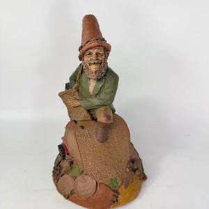 May include: A whimsical figurine of a gnome wearing a brown hat and green jacket, sitting on a heart-shaped base with coins and leaves. The gnome is holding a scroll in his hands.