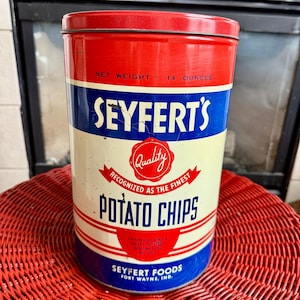 May include: A cylindrical metal Seyfert's Potato Chips tin with a red, white, and blue color scheme. The label reads NET WEIGHT 14 OUNCES, SEYFERT'S, Quality RECOGNIZED AS THE FINEST, POTATO CHIPS, INGREDIENTS SELECT POTATOES VEGETABLE OIL AND SALT, SEYFERT FOODS FORT WAYNE, IND.