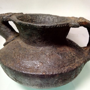 May include: A dark brown ceramic pot with a wide mouth and two handles. The pot has a rough, textured surface and appears to be very old.