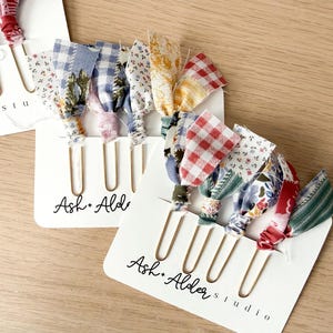 May include: A set of gold-toned metal paper clips attached to white display cards labeled Ash + Alder studio. Each clip features a tied fabric scrap in various patterns including red and blue gingham, floral prints, and stripes in multiple colors.