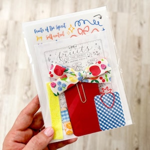 May include: A stationery set in a clear plastic sleeve featuring a fabric bow with a fruit pattern, a gold paperclip, a solid red card, a blue and white gingham card with a red strawberry-shaped paperclip, and a yellow strip of paper.