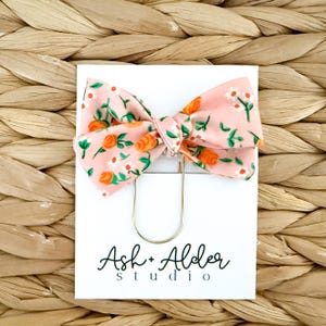 May include: A pink floral bow tie with orange and white flower accents, attached to a gold paper clip. The bow tie is on a white card with the text "Ash + Alder studio" in black script.