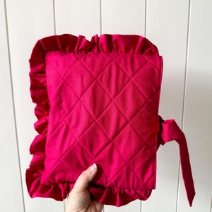 May include: A vibrant fuchsia-colored quilted pillow with a ruffled edge and a matching tie. The pillow features a diamond pattern and is made of a soft fabric. This decorative pillow adds a pop of color.