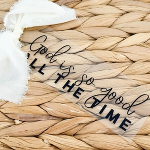 May include: A clear, rectangular tag with the words "God is so good all the time" in black script and block letters. A white ribbon with frayed edges is tied to the tag, set against a woven, light brown background.