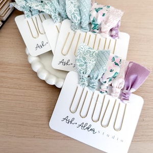 May include: Hair accessories displayed on white cards with gold-colored hair pins. The cards have fabric bows in various floral patterns and solid colors. The text "Ash. Alder studio" is printed on the cards.