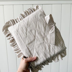 May include: A beige and white striped quilted pillow with ruffled edges. The pillow has a diamond pattern and is tied with a matching fabric bow. The pillow is approximately 12 inches square.