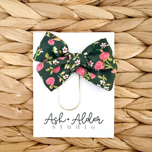 May include: A dark green fabric bow with a floral pattern of pink roses and white flowers. The bow is attached to a white card with the text "Ash + Alder studio" in black script. A gold-colored paper clip holds the bow in place.