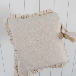 May include: A square cream-colored quilted pillow featuring a delicate white floral pattern. The pillow is edged with a ruffled fabric border and includes a fabric tie on one side, providing a soft and textured appearance to the square cushion.