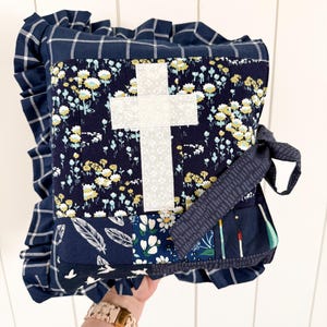 May include: A handmade, quilted pillow featuring a white cross applique on a navy floral fabric. The pillow has a ruffled edge and a navy plaid top. A navy ribbon is tied to the side. The pillow is a religious item.
