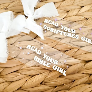 May include: Clear acrylic bookmark with the text "READ YOUR SCRIPTURES GIRL" and "READ YOUR BIBLE GIRL" in white, accented with purple star shapes. A white ribbon is attached to the bookmark. The background is a woven, light brown material.