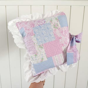 May include: A square patchwork pillow featuring a variety of light pink and blue floral, polka dot, and gingham fabric squares. The pillow is edged with a wide white eyelet lace ruffle and includes a matching fabric bow tied on the side.