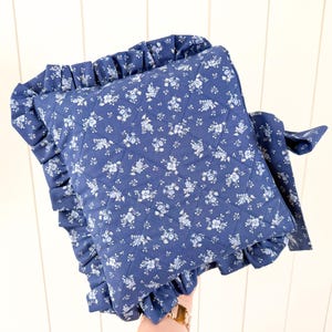 May include: A navy blue quilted pillow with a white floral pattern and a ruffled edge. The pillow has a square shape and a matching fabric tie. The pillow is approximately 14 inches square.