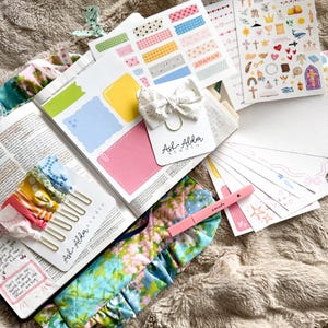 May include: An open book with colorful sticky notes, decorative stickers, and a bow-shaped paper clip. The book is surrounded by a floral fabric cover, bookmarks, and a pink pen with the word "smile". The items are arranged on a soft, textured surface.