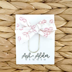 May include: A white fabric bow hair clip with a pink bow pattern, attached to a white card with the text "Ash + Alder studio" in black script. The clip is gold-colored.