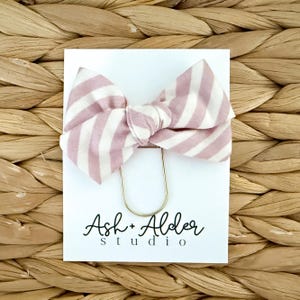 May include: A striped pink and white bow hair accessory is attached to a white card with a gold-colored metal clip. The card features the text "Ash + Alder studio" in a handwritten font. The background is a woven texture.