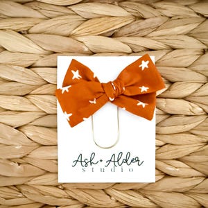 May include: A burnt orange fabric bow with white star patterns, attached to a white card with gold paper clip. The card features the text "Ash + Alder studio" in a handwritten style font. The background is a woven wicker texture.