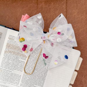 May include: A white mesh bow with colorful polka dots and embroidered accents, clipped to a book with a gold paperclip. The book is open to a page with text and a tab labeled "Exodus."