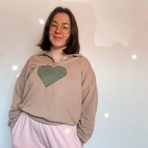 Peut inclure: Un chemisier beige à manches longues avec un motif de cœur vert. Le chemisier a un col boutonné et une coupe décontractée.