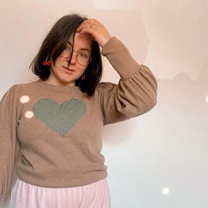 Peut inclure: Un pull marron avec un patch en forme de cœur vert et blanc sur le devant. Le pull a des manches longues et un col rond.