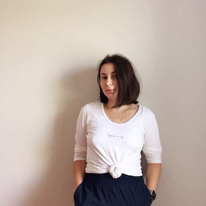 Peut inclure: Une femme portant un chemisier blanc à manches longues avec le texte "Supreme in my way" brodé sur le devant, noué à la taille, et un pantalon bleu foncé.