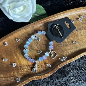 May include: A beaded bracelet with iridescent blue and purple beads, silver charms of a wolf and a heart, and a black guitar pick with gold text. The bracelet is on a wooden tray with decorative crystals and a white rose.
