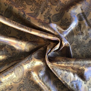 May include: Gold fabric with a paisley pattern in black. The fabric is folded and draped, showing the texture and sheen of the material.