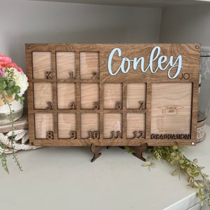 May include: A wooden photo frame with 12 slots for pictures, labeled with numbers from K to 12. The frame has a graduation cap and the text "Class of 2037" and "Graduation" on it. The name "Conley JO" is written on the top of the frame.