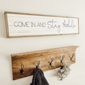 May include: A wooden coat rack with four hooks and the text "Come in and Stay Awhile" on a sign above it.