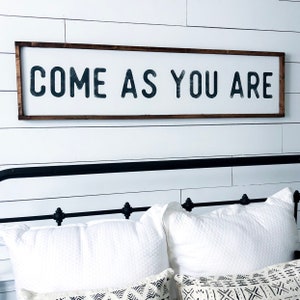 May include: A wooden sign with a dark brown frame and white background. The sign reads "COME AS YOU ARE" in black lettering.