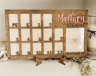 May include: A wooden photo frame with 12 spaces for pictures, numbered from PK to 12. The frame is brown with a light wood interior. The frame has a graduation cap and the text "Class of 2023" and "Graduation". The name "Mallory Leeann" is written in pink letters on the frame.