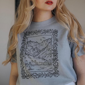 May include: A light blue t-shirt featuring a black and white mountain landscape design within a floral frame. The design includes a sailboat on a lake and birds in the sky. The shirt is a soft, casual style.
