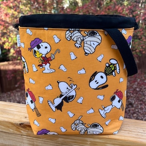 May include: A bright orange drawstring bag with a black top and black straps. The bag is decorated with a repeating pattern of Snoopy dressed in various Halloween costumes, including a pirate, a mummy, a vampire, and a skeleton. There are also small white ghost images scattered throughout the pattern.