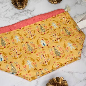 Könnte beinhalten: Eine gelbe Kordeltasche mit rosa-weißem Besatz. Die Tasche hat einen weißen Baumwollgriff und zeigt ein sich wiederholendes Muster aus Lebkuchenmännern, Zuckerstangen und Weihnachtsbäumen.