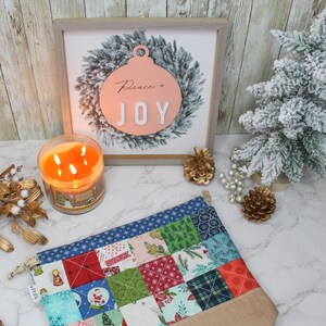 Può includere: Una pochette in tessuto trapuntato con un design a patchwork in tonalità di blu, verde, rosso e bianco. La pochette è appoggiata su una superficie bianca con una candela accesa, un cartello di legno con un ornamento rosa che dice "Peace & Joy", e un piccolo albero artificiale ricoperto di neve.