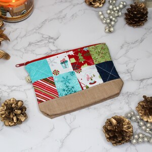 Puede incluir: Un estuche acolchado con temática navideña con cremallera roja. El diseño de patchwork presenta cuadrados con cajas de regalo, copos de nieve y árboles de Navidad. La parte inferior es de tela beige lisa.