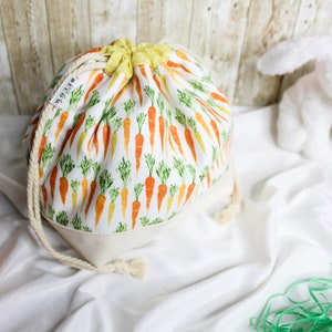 May include: A white drawstring bag with a carrot print. The bag is filled with green and orange carrots. The bag is sitting on a white surface with a white stuffed bunny and a green Easter grass.