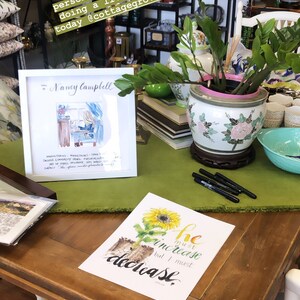 May include: A white frame with a watercolor painting of a woman sitting at a desk. The painting is titled "Nancy Campbell". The frame is sitting on a wooden table with a green tablecloth. There is a potted plant with green leaves and a white and pink pot on the table. There is also a black and white print with a sunflower and the text "He must increase but I must decrease" on the table.