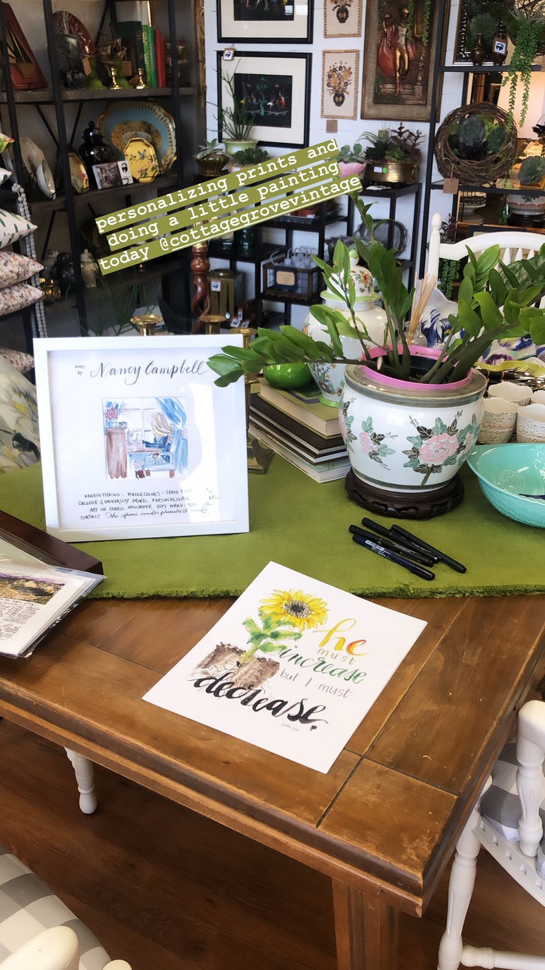 May include: A white frame with a watercolor painting of a woman sitting at a desk. The painting is titled "Nancy Campbell". The frame is sitting on a wooden table with a green tablecloth. There is a potted plant with green leaves and a white and pink pot on the table. There is also a black and white print with a sunflower and the text "He must increase but I must decrease" on the table.