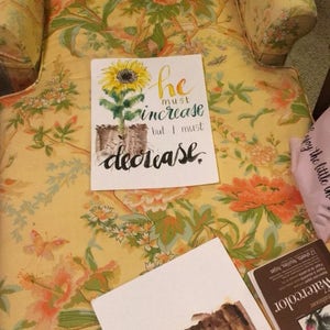 May include: A watercolor painting of a sunflower with the text "he must increase but I must decrease." The painting is on a white background and is sitting on a floral patterned chair.