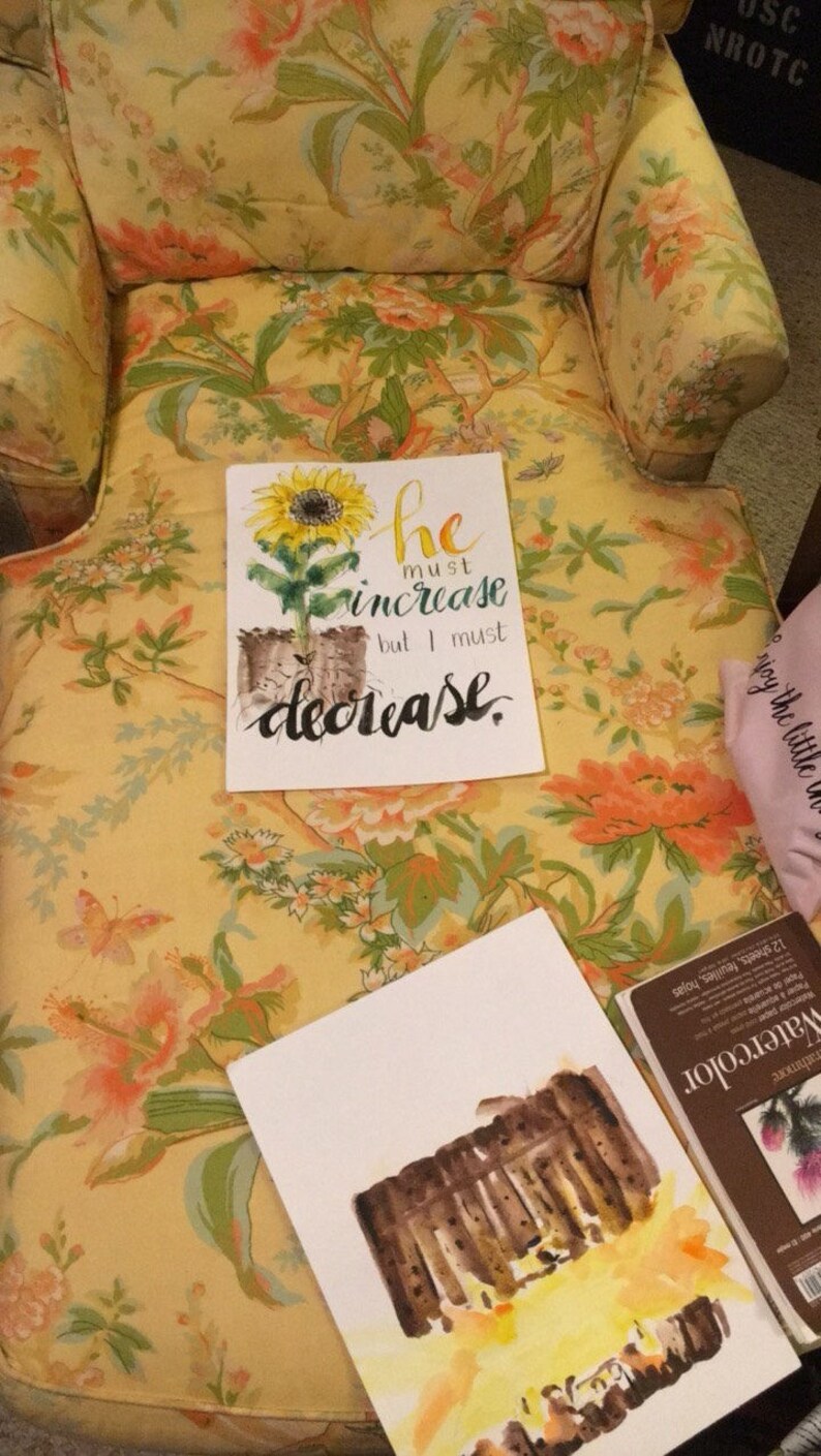 May include: A watercolor painting of a sunflower with the text "he must increase but I must decrease." The painting is on a white background and is sitting on a floral patterned chair.
