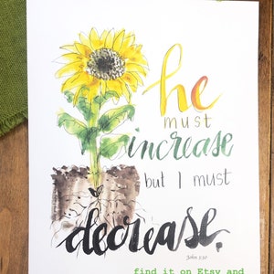 May include: A watercolor painting of a sunflower with the text "he must increase but I must decrease" and the scripture reference "John 3:30". The painting is on a white background and has a green and brown watercolor wash around the sunflower.